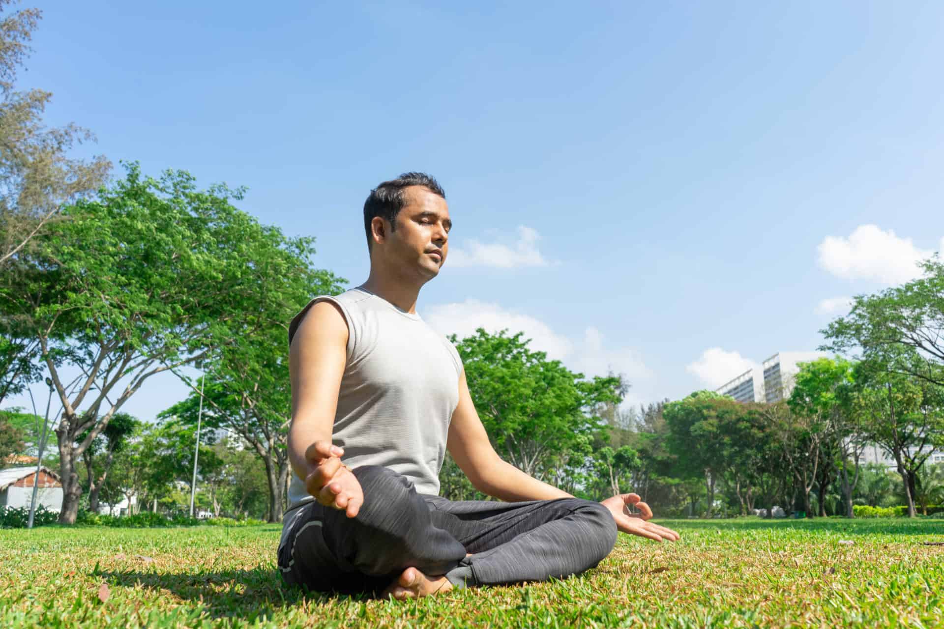 Indian man meditating in lotus pose outdoors on summer lawn with trees in background. Outdoor yoga concept.