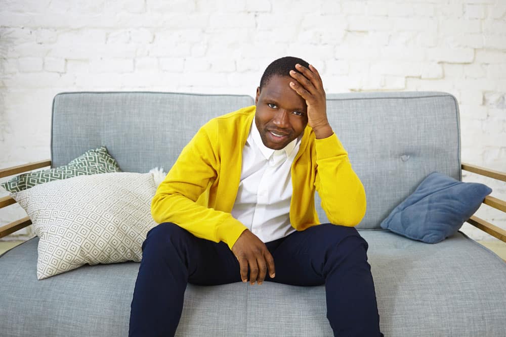 portrait-sad-unhappy-african-male-yellow-cardigan-sitting-couch-with-decorative-pillows-keeping-hand-head-feeling-nervous-while-watching-tv-football-match-having-worried-expression