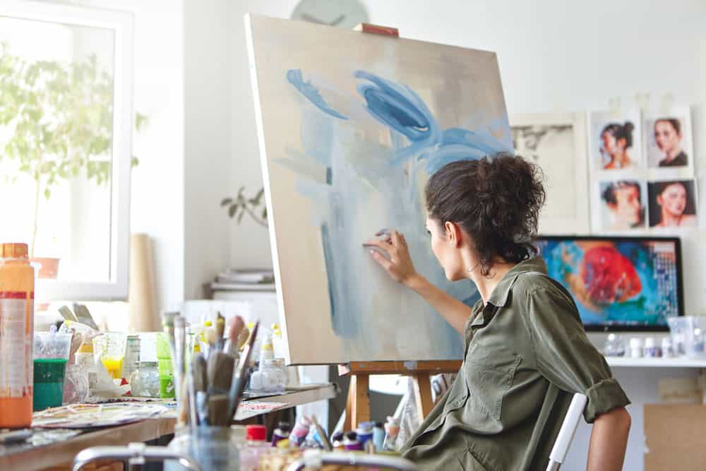 art-creativity-hobby-job-creative-occupation-concept-rear-view-busy-female-artist-sitting-chair-front-easel-painting-with-fingers-using-white-blue-oil-acrylic-paint